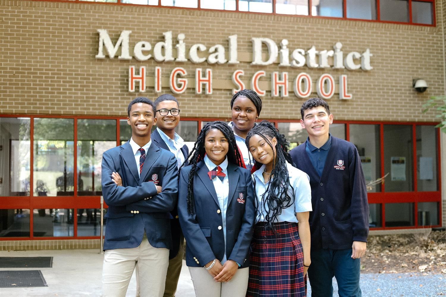 A group of six students, smiling and wearing school uniforms, stand in front of the "Medical District High School" building entrance.