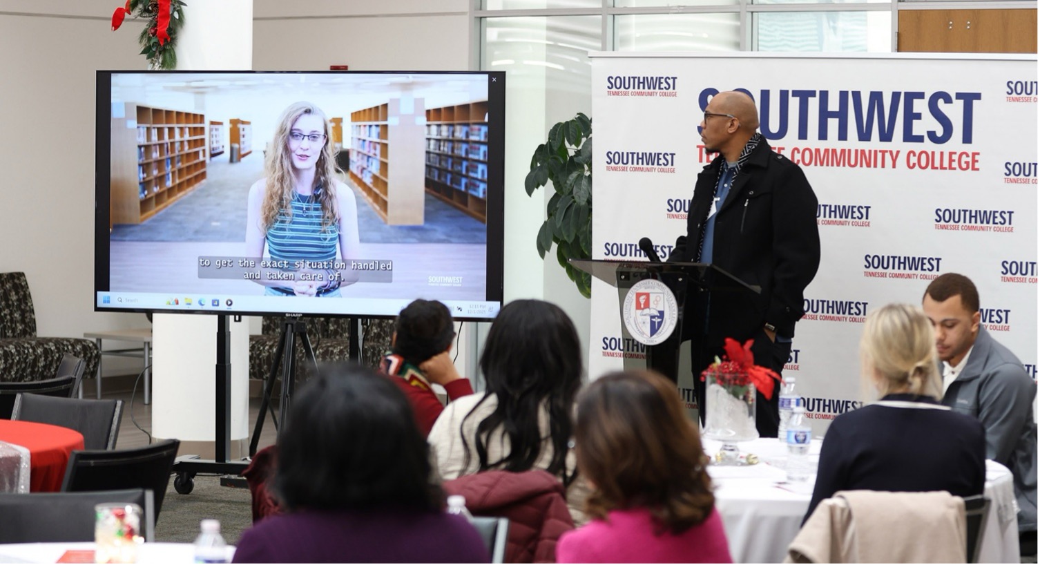 Speaker at a Southwest Tennessee Community College podium addresses guests at round tables; a screen shows a subtitled library video; holiday décor with red bows and greenery.