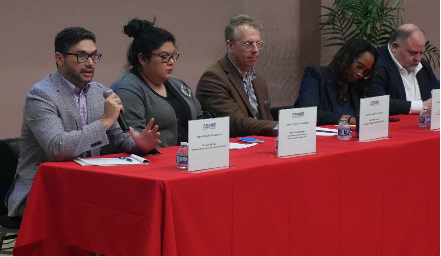 A panel of five people sit behind a red tablecloth, engaged in discussion. One man speaks into a microphone. Nameplates and water bottles are visible.