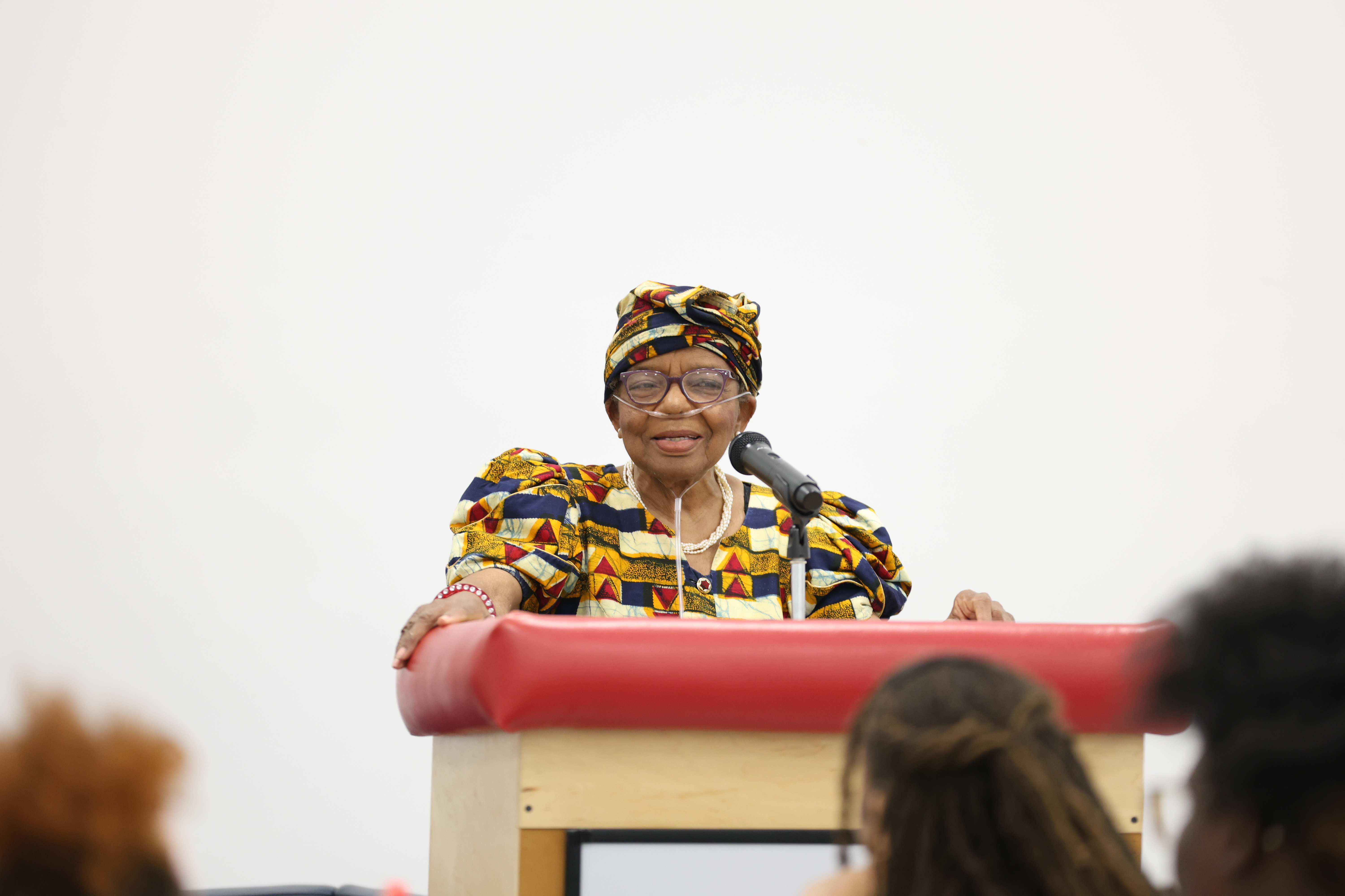 A speaker wearing a brightly patterned outfit stands behind a red topped podium, addressing an audience in a brightly lit room.