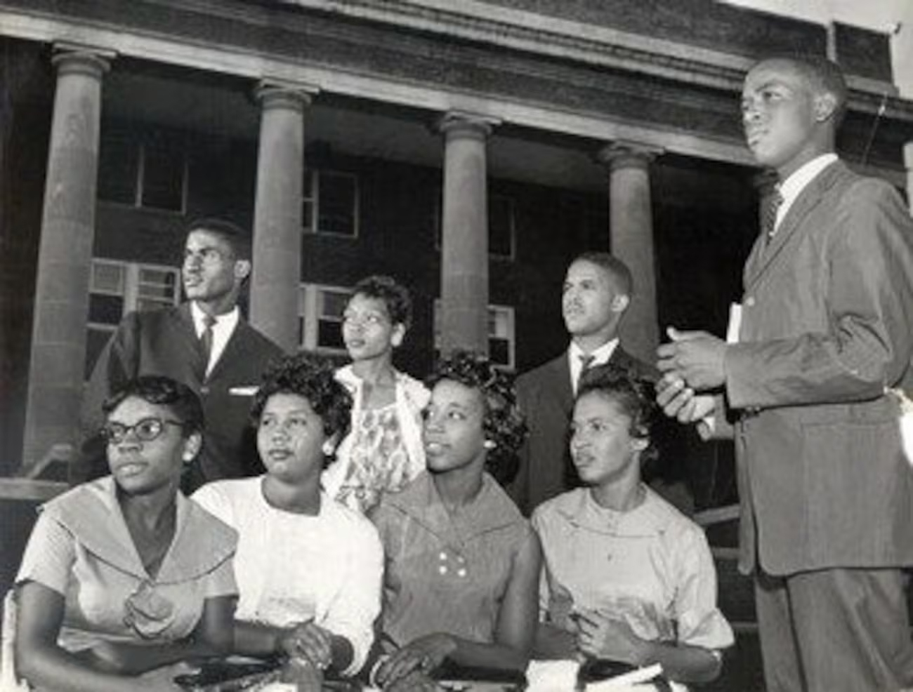 A historic black and white photo of eight young African American students posing together in front of a large brick building with tall columns. The group sits and stands in two rows, dressed in mid 20th century attire.