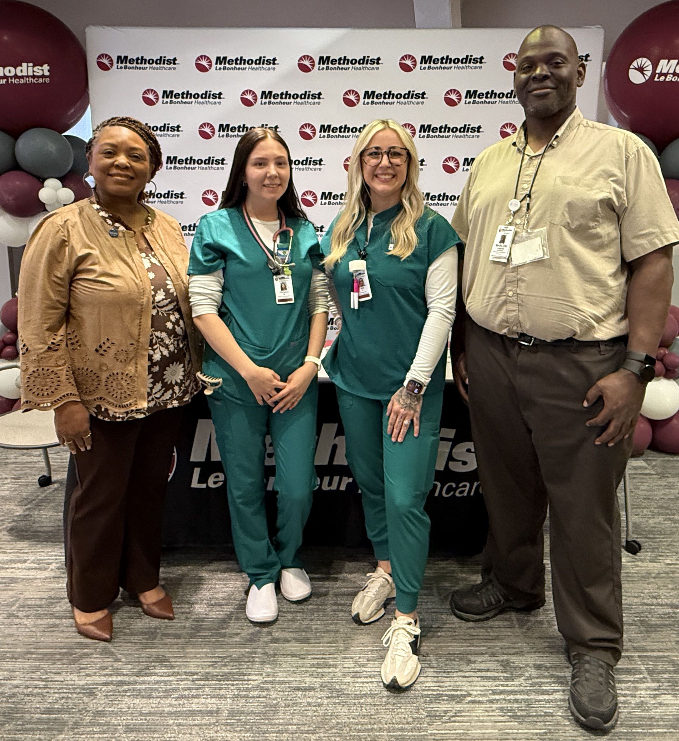 Four people stand posing for a photo in front of a backdrop printed with multiple Methodist Le Bonheur Healthcare logos. Two of the individuals are wearing teal medical scrubs, and the other two are dressed in business or casual attire. A large balloon display stands behind them on both sides. A screen to the left shows part of an event welcome slide, and a sign to the right reads “Improving Every Life We Touch.”